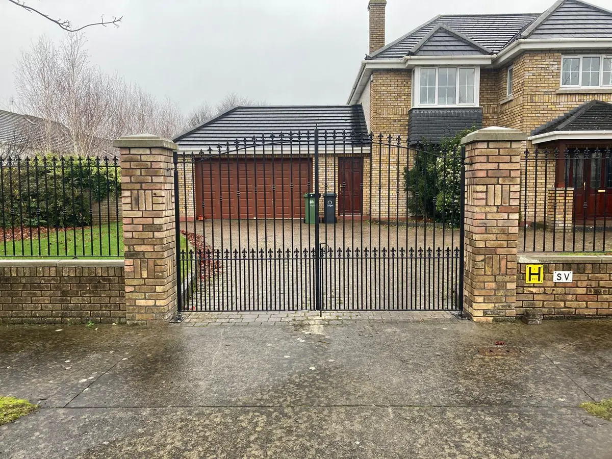 A pair of arched wrought iron style driveway gates in black steel with fleur de lis finials and dog bars, hung on brick piers, made and fitted by our gate maker