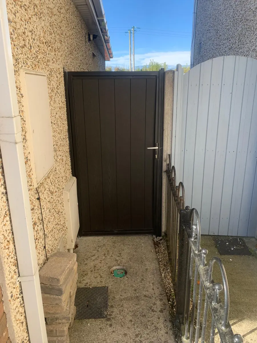 A brown composite infill metal side gate in a black steel frame fitted beside a pebble dashed house