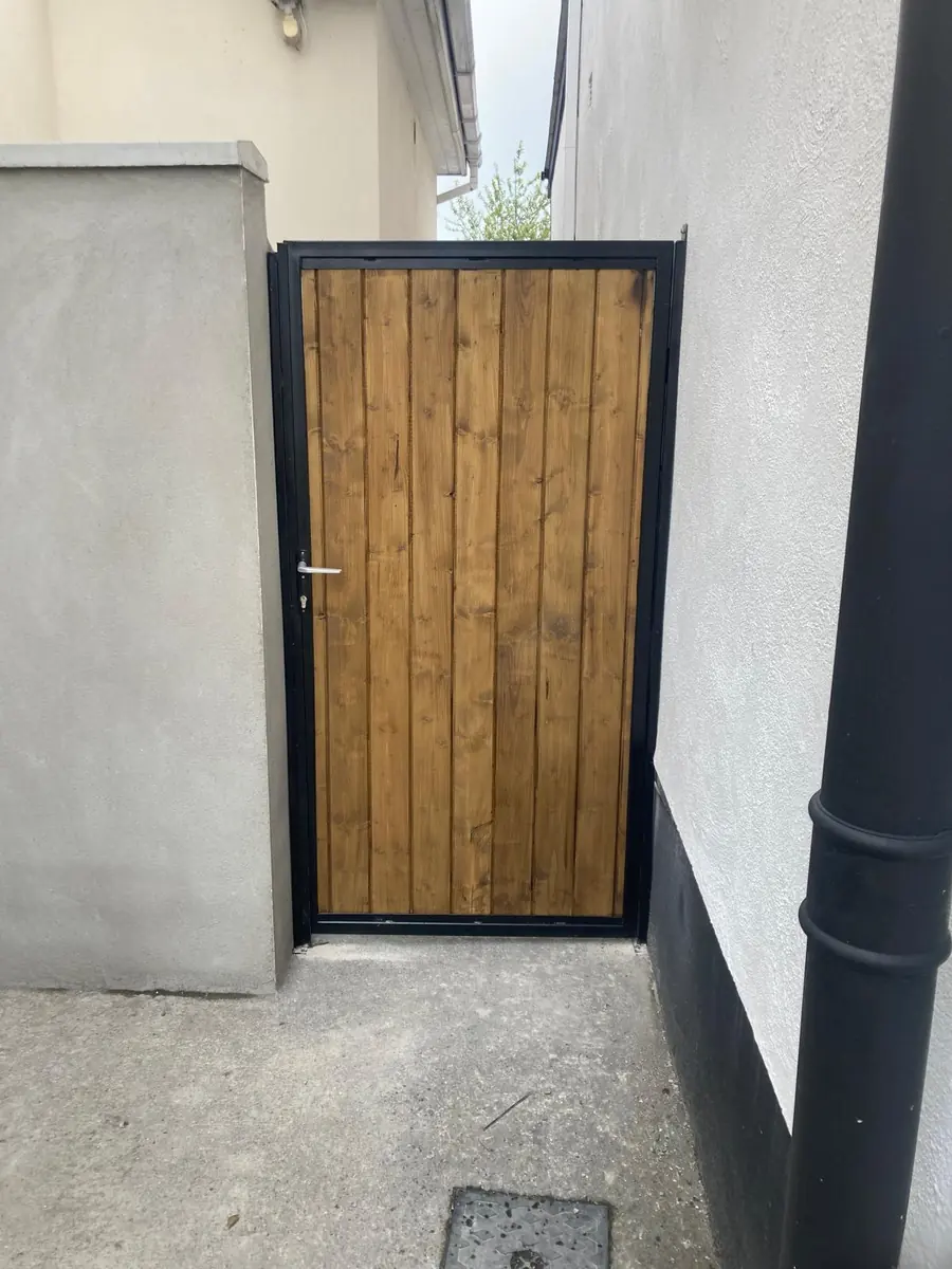 A made to measure side gate with a black steel frame and vertical timber board infill beside a rendered house